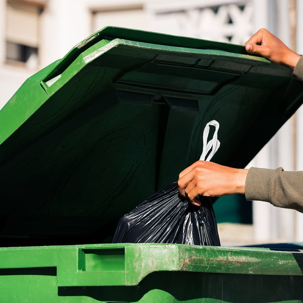 Man throwing out black eco-friendly recyclable trash bag in to big plastic green garbage container